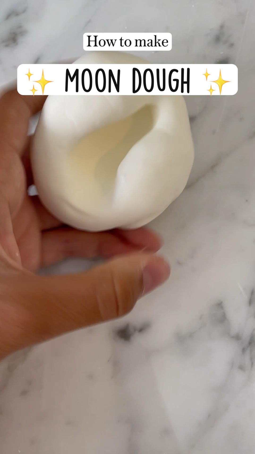 A person's hand holds a smooth, pale white blob of moon dough with a finger indentation, resting on a white marble.