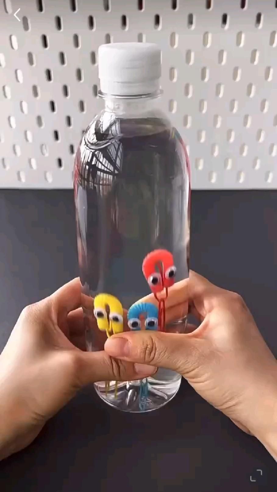 Hands holding a clear plastic water bottle filled with water containing small colorful figures made from paperclips and.