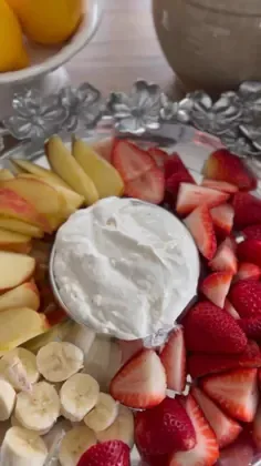 Silver bowl of creamy white fruit dip surrounded by fresh strawberries, banana slices, and apple slices on a floral platter.