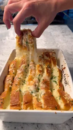 Hand pulling apart a slice of cheesy garlic bread with melted cheese and fresh parsley from a white baking dish