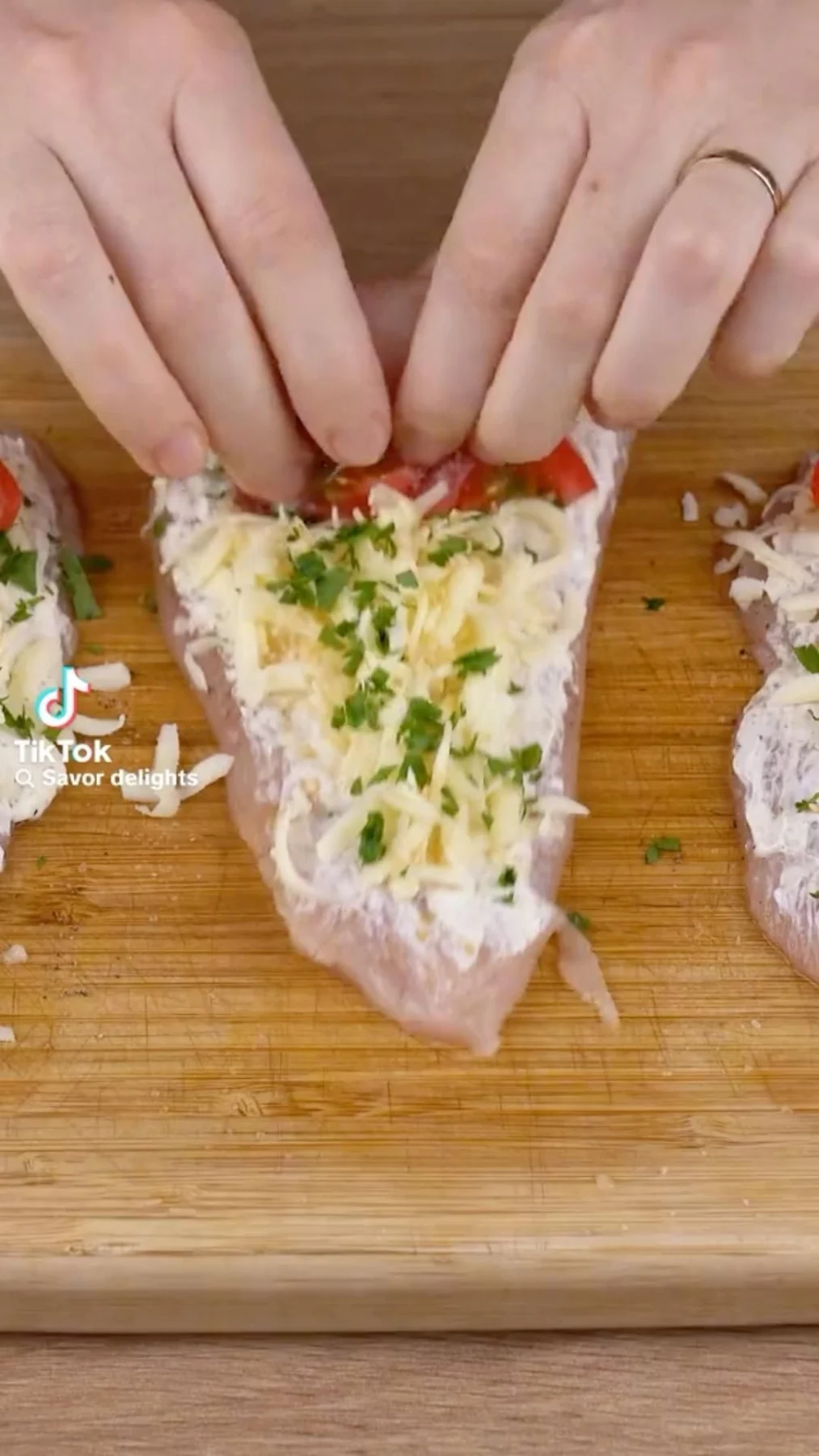 Hands spreading cream cheese on sliced chicken breasts with shredded cheese, parsley, and cherry tomatoes nearby.