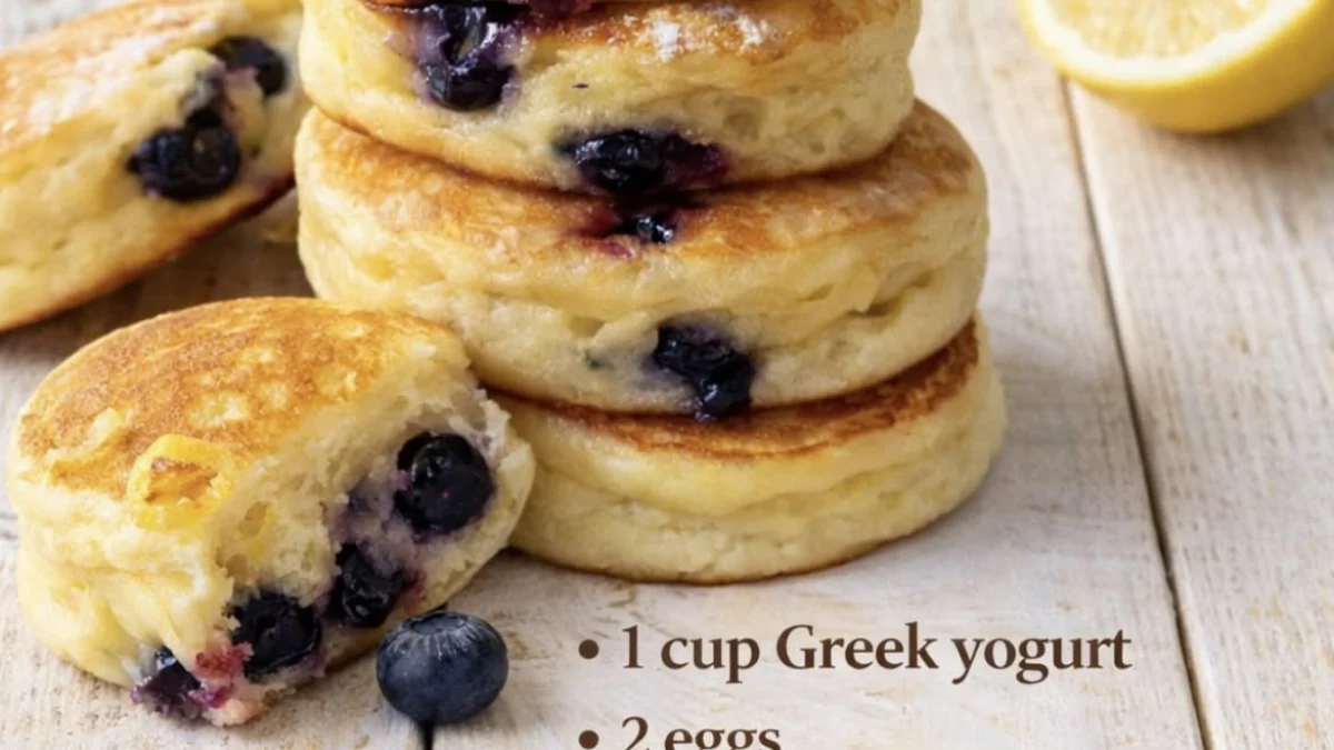 A stack of golden-brown pancake bites with blueberries and lemon zest on a wooden surface with lemons.
