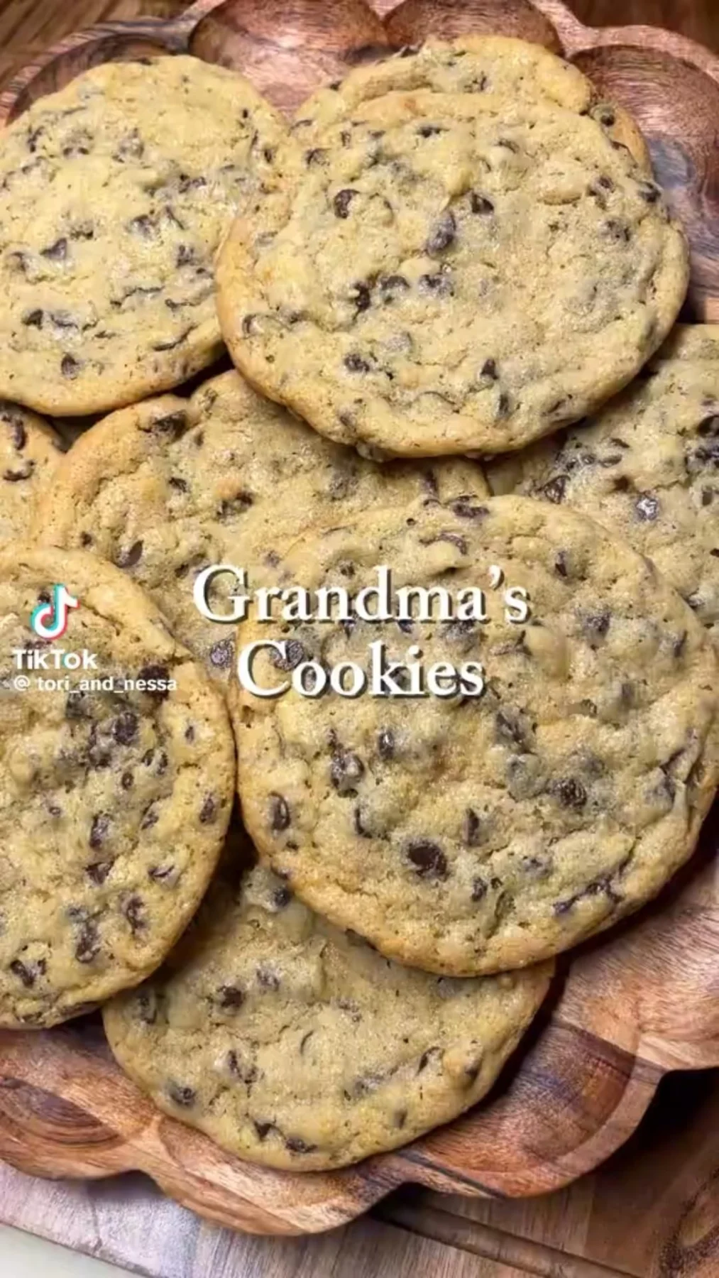 Several soft golden-brown chocolate chip cookies stacked on a textured wooden serving board.