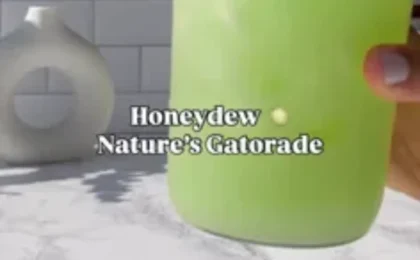 A tall glass of pale green honeydew drink with a slice of honeydew on the rim, next to fresh mint leaves.