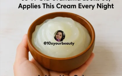 A 63-year-old woman with luminous bare skin holding a small glass jar of rich ivory cream near her smiling face