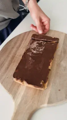 Hand holding a rectangular protein bar coated in glossy dark chocolate on a wooden cutting board