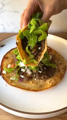 A hand holding up a smash burger taco with ground beef, lettuce, tomatoes, red onion, cotija cheese, and sauce on a ceramic.