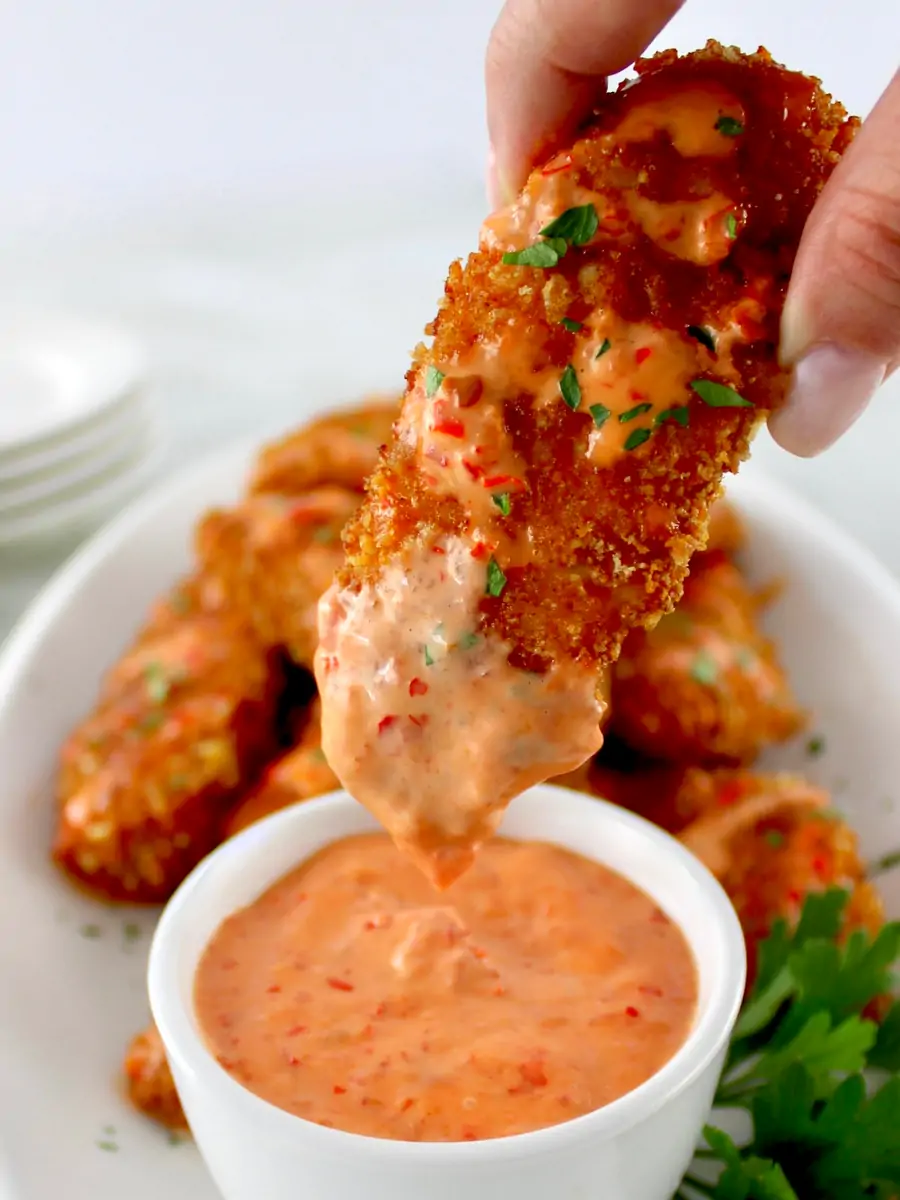 Hand holding a crispy bang bang chicken tender dipped in creamy orange sauce over a bowl of sauce