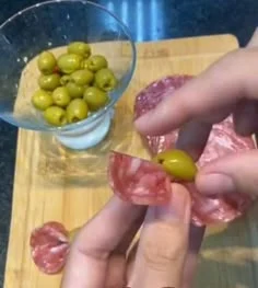 Hands placing a green olive on a folded salami slice on a wooden board