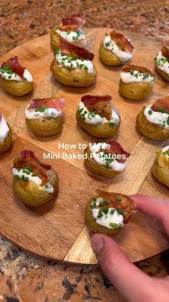 Mini baked potatoes topped with sour cream, bacon bits, and chopped chives on a wooden board