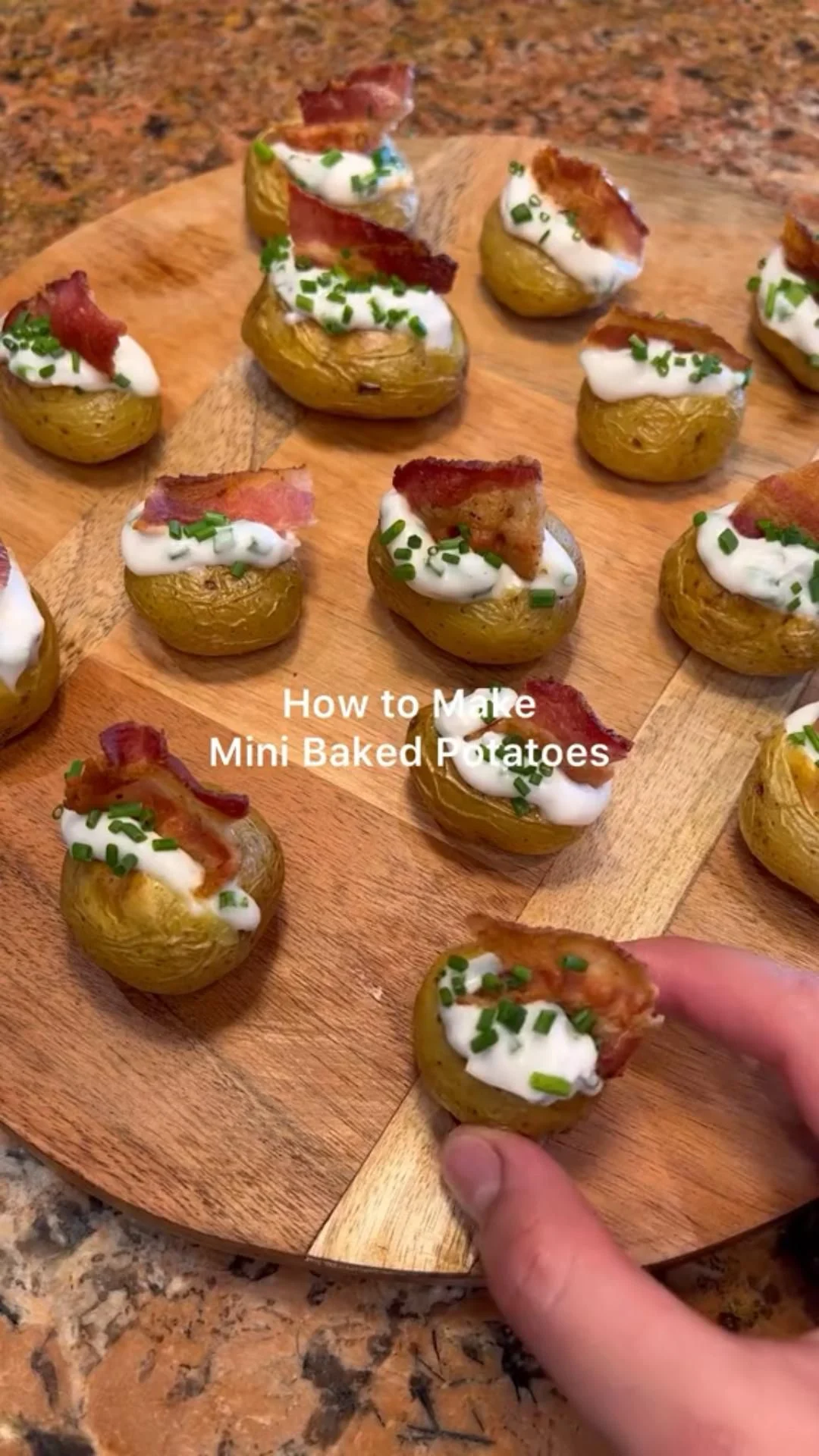 Mini baked potatoes topped with sour cream, bacon bits, and chopped chives on a wooden board