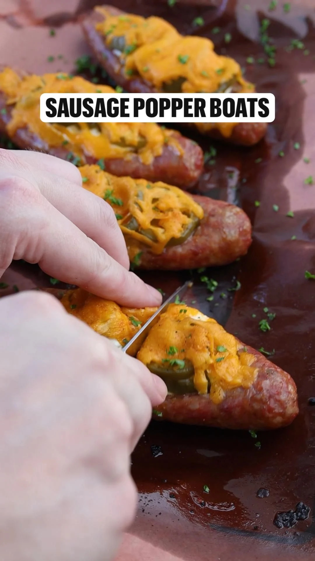 Hands spreading cheese and peppers into split smoked sausage boats on a parchment-lined sheet