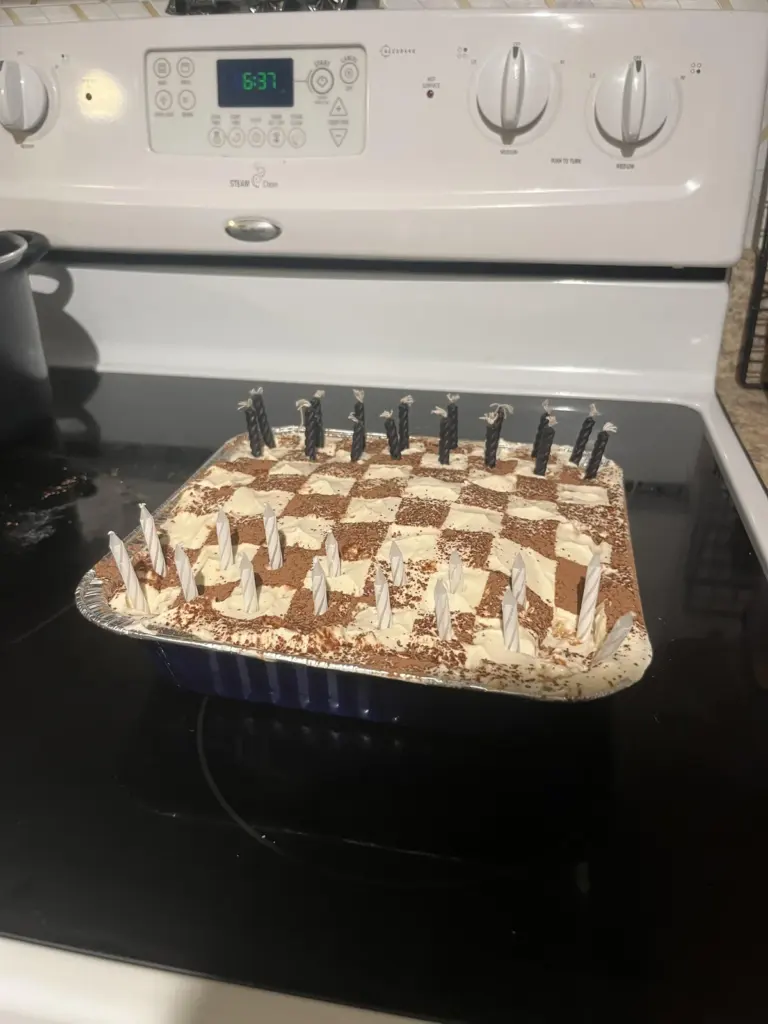 A rectangular tiramisu topped with a chess board pattern of alternating dark cocoa powder and light cream squares, with black birthday candles placed on top.