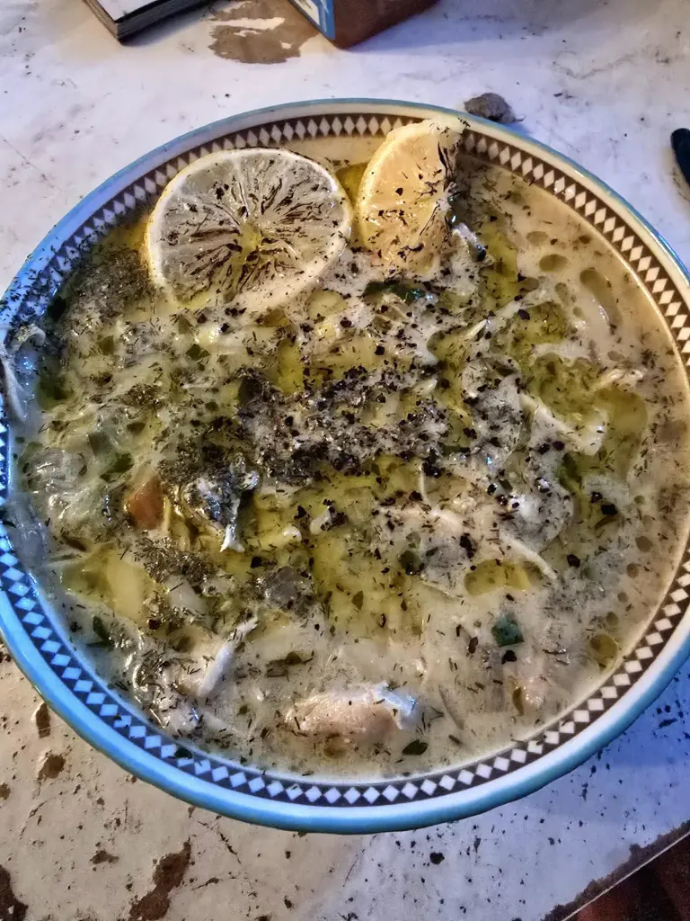 A bowl of creamy Greek avgolemono soup with shredded chicken, rice, and a sprinkle of fresh parsley, with a lemon wedge on the side.