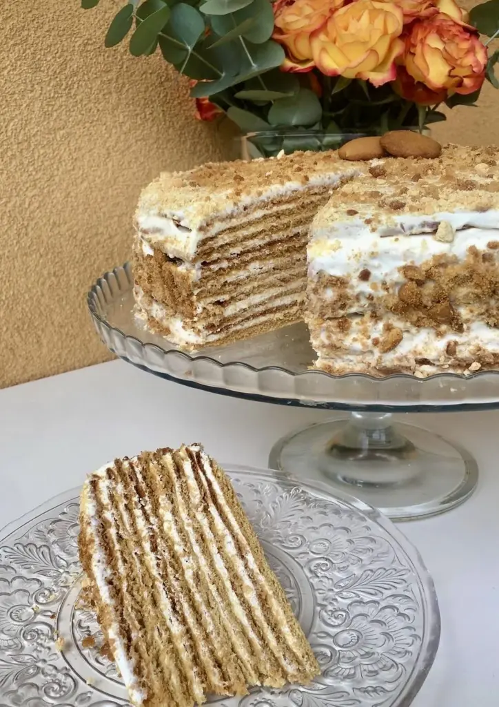 A slice of Russian honey cake (medovik) showing many thin golden-brown layers separated by white sour cream frosting, with crumbs on top.